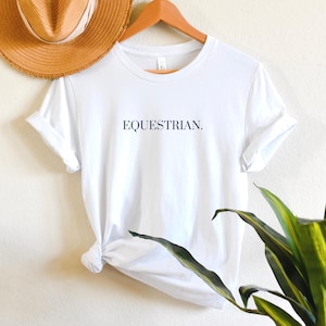 K&ouml;nnte beinhalten: Wei&szlig;es T-Shirt mit dem Text "EQUESTRIAN." in dunkelblau auf der Vorderseite.