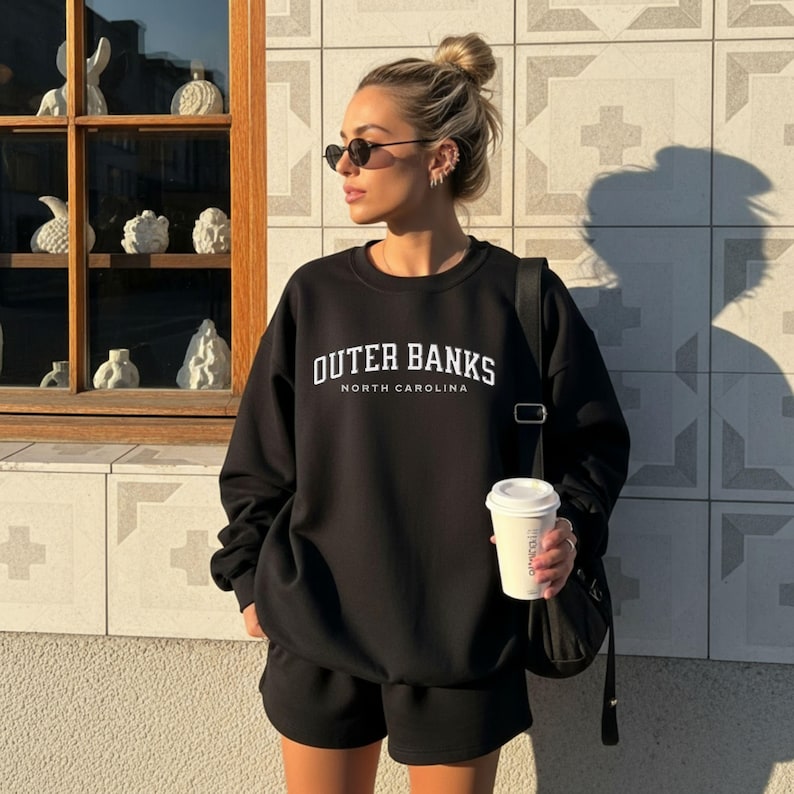 May include: Black sweatshirt and shorts set with the text "OUTER BANKS NORTH CAROLINA" in white. The person is wearing sunglasses and holding a coffee cup. The background includes a window with decorative objects and a tiled wall.