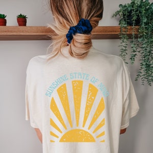May include: Cream-colored t-shirt with a yellow sunburst design and the text "SUNSHINE STATE OF MIND" in light blue. The shirt is a casual style, perfect for everyday wear. The back of the shirt is visible.