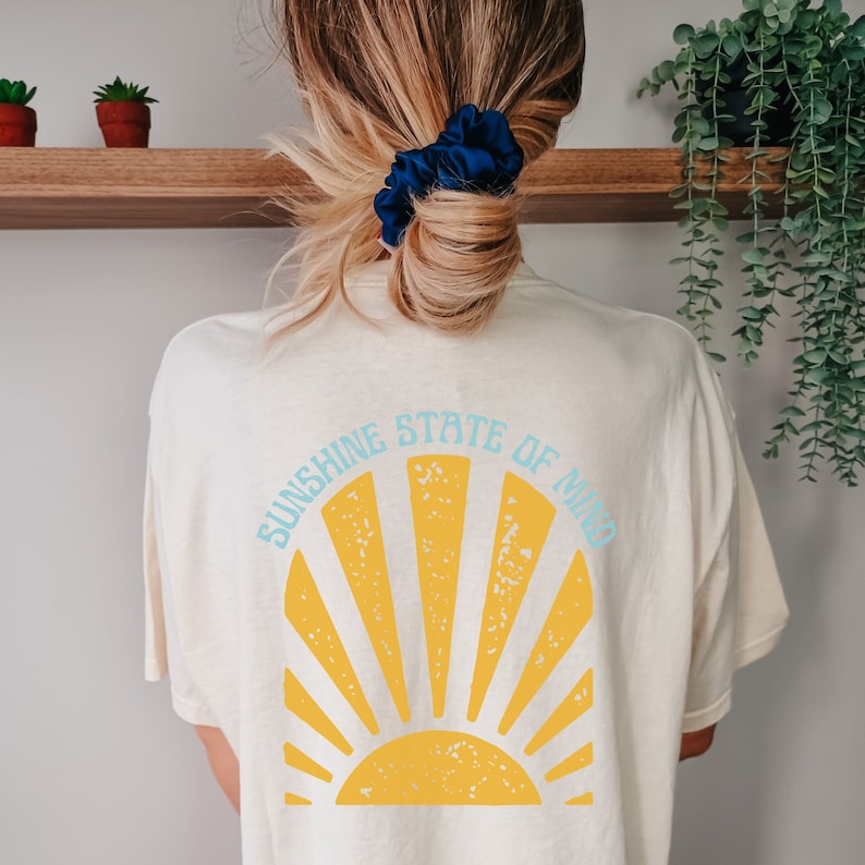 May include: Cream-colored t-shirt with a yellow sunburst design and the text "SUNSHINE STATE OF MIND" in light blue. The shirt is a casual style, perfect for everyday wear. The back of the shirt is visible.