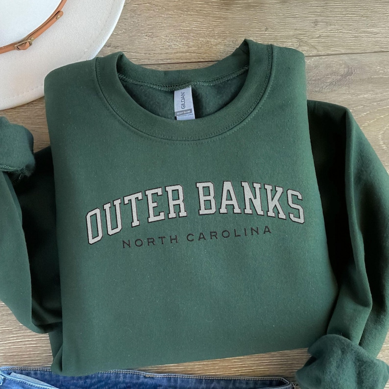 May include: A dark green sweatshirt with the words "OUTER BANKS" in a white, block font, arched across the chest, with "NORTH CAROLINA" below. The sweatshirt is folded on a wooden surface, with a white hat visible in the background.
