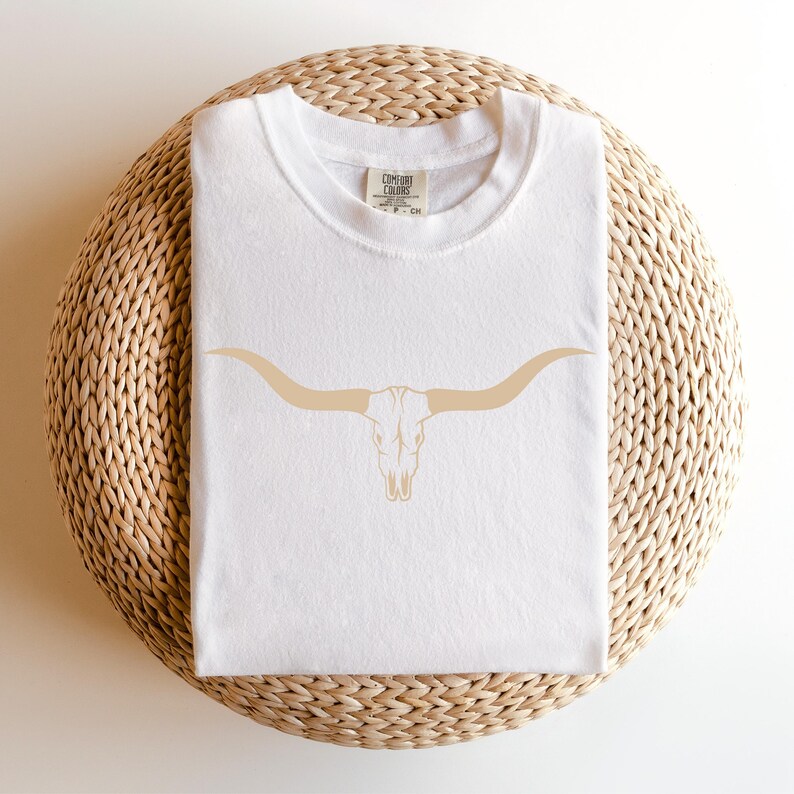 Op de afbeelding: Wit T-shirt met een bruine omtrek van een longhorn schedel met hoorns. Het shirt is gevouwen en ligt op een geweven stro mat.
