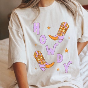 Puede incluir: Una camiseta blanca con la palabra "HOWDY" impresa en morado con dos botas de vaquero naranja y morado con dise&ntilde;os florales.