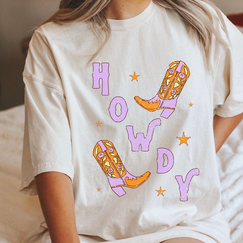 Puede incluir: Una camiseta blanca con la palabra "HOWDY" impresa en morado con dos botas de vaquero naranja y morado con dise&ntilde;os florales.