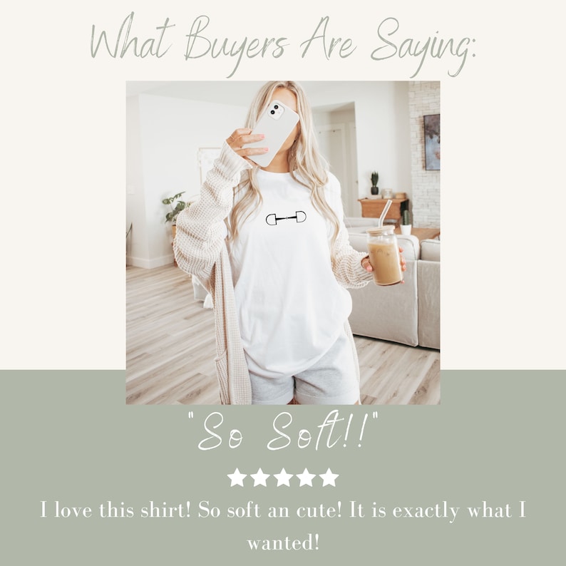May include: A woman wearing a white t-shirt with a black horse bit design. The shirt has the words "So Soft!" written on it. The woman is holding a phone in her right hand and a glass in her left hand. The background is a light-colored room.