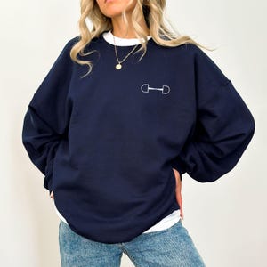 May include: A navy blue sweatshirt with a white horse bit design on the front. The sweatshirt has long sleeves and a relaxed fit.