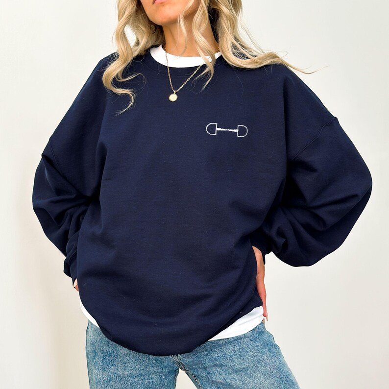 May include: A navy blue sweatshirt with a white horse bit design on the front. The sweatshirt has long sleeves and a relaxed fit.