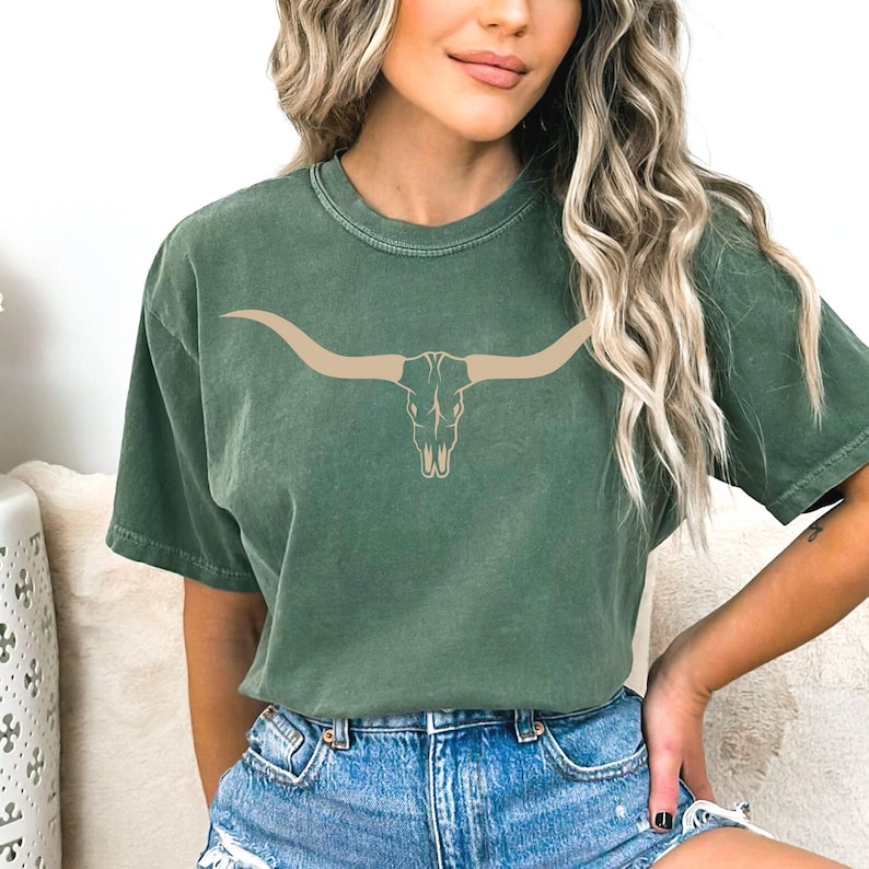 Op de afbeelding: Een groen T-shirt met korte mouwen met een tanne stieren schedel grafische afbeelding. De schedel heeft grote hoorns.