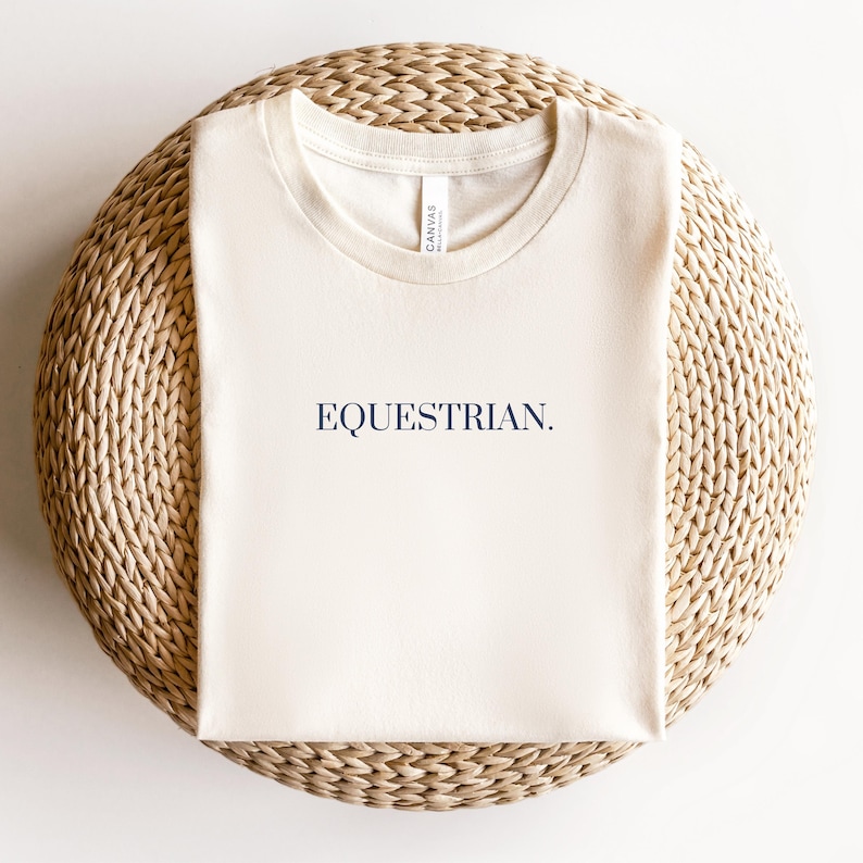 K&ouml;nnte beinhalten: Ein cremefarbenes T-Shirt mit dem Schriftzug "EQUESTRIAN." in Marineblau auf der Vorderseite. Das Shirt liegt auf einer geflochtenen Strohh&uuml;tte.