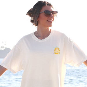 May include: Cream-colored t-shirt with a round neck and short sleeves. The shirt features a small graphic of a rising sun with the text "Sunshine State of Mind" in light blue. The shirt is made of a soft, comfortable material.