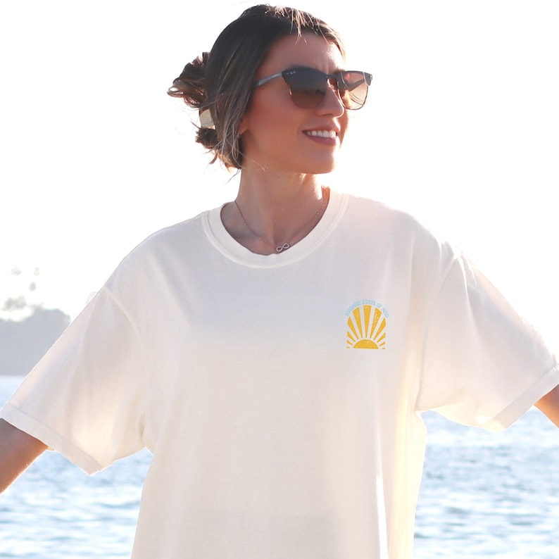 May include: Cream-colored t-shirt with a round neck and short sleeves. The shirt features a small graphic of a rising sun with the text "Sunshine State of Mind" in light blue. The shirt is made of a soft, comfortable material.