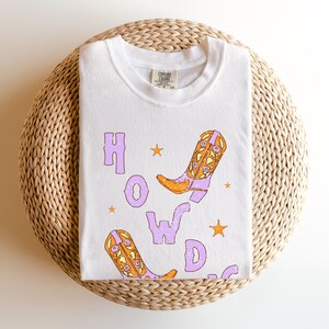 Puede incluir: Camiseta blanca con un gr&aacute;fico morado y naranja de botas de vaquero y la palabra "HOWDY" en una fuente desgastada.