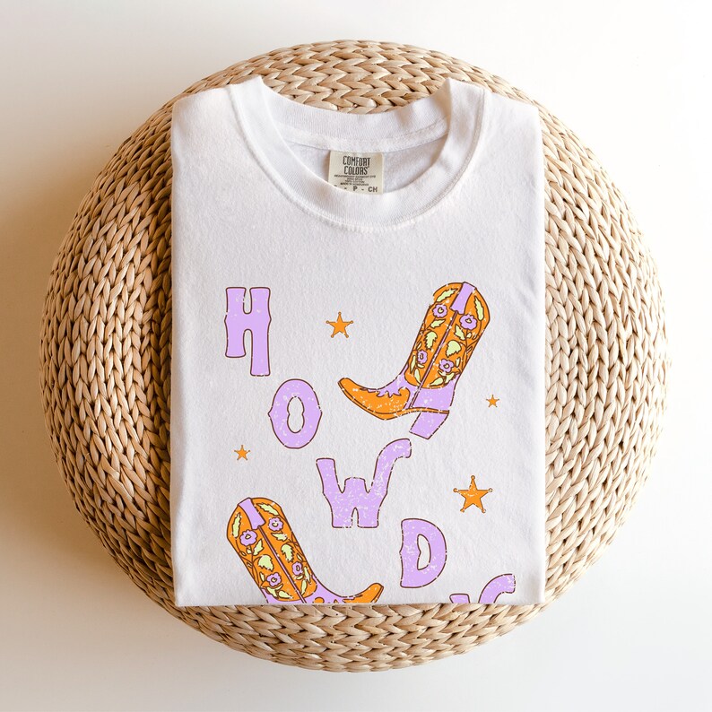 Puede incluir: Camiseta blanca con un gr&aacute;fico morado y naranja de botas de vaquero y la palabra "HOWDY" en una fuente desgastada.