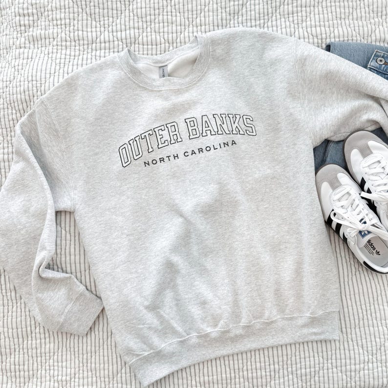 May include: A light gray sweatshirt with the words "OUTER BANKS NORTH CAROLINA" printed in black. The sweatshirt is paired with blue jeans and white sneakers with black stripes.
