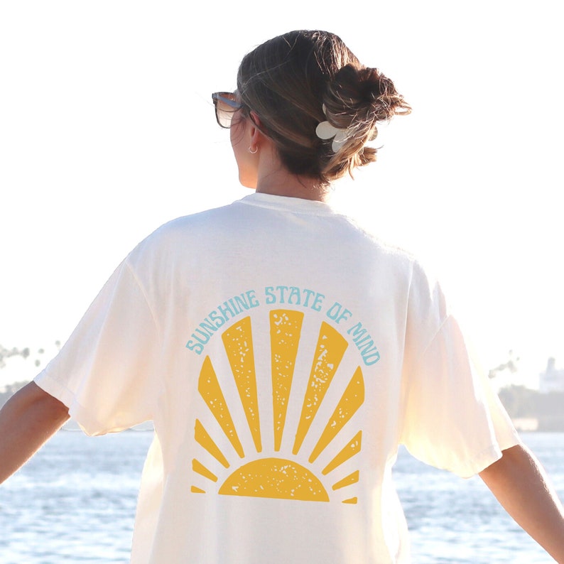 May include: Cream-colored t-shirt with a yellow sunburst design and the text "SUNSHINE STATE OF MIND" in light blue. The shirt is visible from the back, with the sun graphic centered. The shirt is a casual wear item.