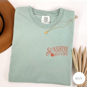 May include: A light blue t-shirt with the text "Sunshine State of Mind" in orange lettering. The shirt has a distressed look and is made of a soft, comfortable fabric.