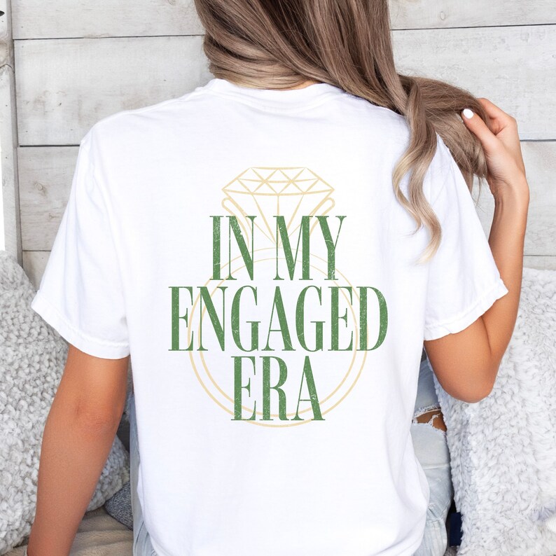 Pu&ograve; includere: Maglietta bianca con una grafica verde di un anello di diamanti e il testo "In My Engaged Era".