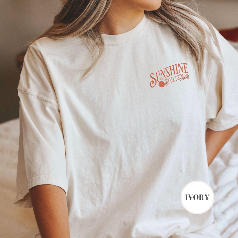 May include: A white oversized t-shirt with the text "Sunshine State of Mind" in orange lettering. The shirt is shown on a model and the color of the shirt is labeled "Ivory".