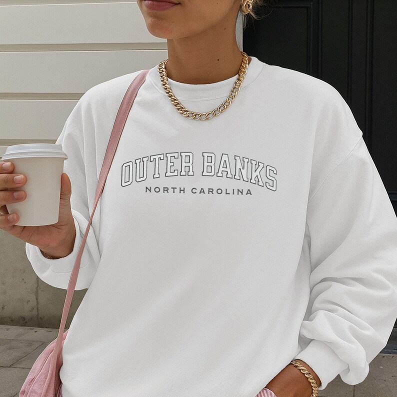 May include: White sweatshirt with the text "OUTER BANKS NORTH CAROLINA" in a black outline. The sweatshirt is paired with a gold chain necklace and a pink shoulder bag. A person is holding a white coffee cup.