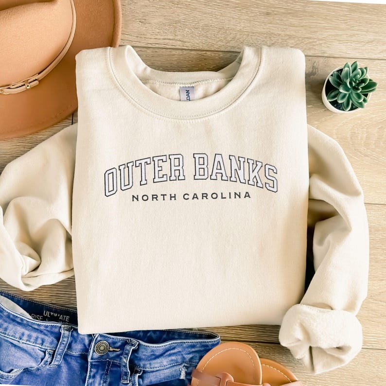 May include: Cream-colored sweatshirt with the text "Outer Banks North Carolina" in black block letters. The sweatshirt is folded on a wooden surface, with a brown hat, blue jeans, and a small succulent plant visible.