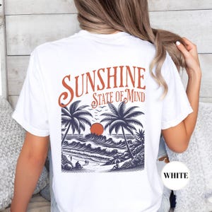 May include: White t-shirt with a graphic of palm trees, waves, and a sunset. The text "Sunshine State of Mind" is printed above the graphic.