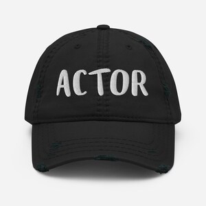 Peut inclure: Casquette de baseball noire délavée avec le mot "ACTOR" brodé en blanc sur le devant. La casquette a une visière incurvée et un aspect usé et vieilli, avec des effilochages visibles sur les bords.
