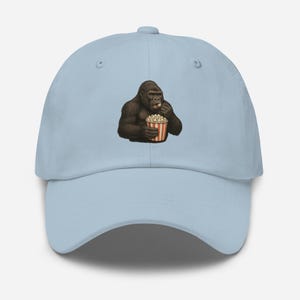 Può includere: Cappellino da baseball azzurro con una grafica di un gorilla che mangia popcorn da un contenitore a strisce rosse e bianche. Il cappellino ha una visiera curva e un bottone. Un accessorio divertente e giocoso.