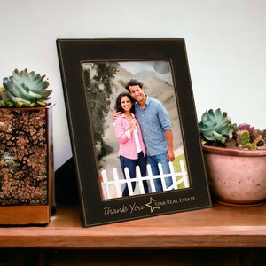 May include: A black leather picture frame with a stitched border and a gold star logo. The frame contains a photo of a couple standing in front of a white picket fence. The text "Thank You" and "STAR REAL ESTATE" is printed on the frame.