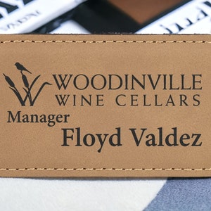 May include: A brown leather name tag with a stitched border. The tag features a logo with the text "Woodinville Wine Cellars" and the word "Manager". The name "Floyd Valdez" is printed below the logo.