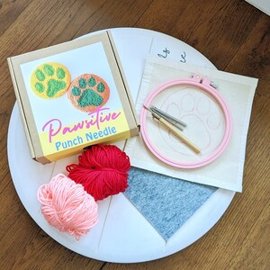 May include: A Pawsitive Punch Needle kit with two paw print designs, pink yarn, red yarn, a wooden embroidery hoop, and a needle. The kit is on a white surface.
