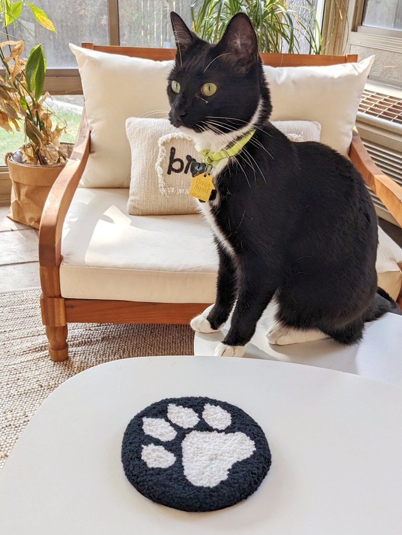 May include: A black and white cat sits on a white chair with a pillow that says "br" on it. The cat is wearing a green collar with a yellow tag that says "Home Again". The cat is looking to the left of the image. A white table with a black and white paw print rug is in the foreground.