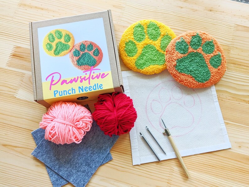 May include: A punch needle kit with the words "Pawsitive Punch Needle" on the box. The kit includes two pre-made paw print shapes, one in yellow and green and the other in orange and green, a piece of white fabric with a paw print outline, two balls of yarn, one pink and one red, two needles, and a piece of gray felt.