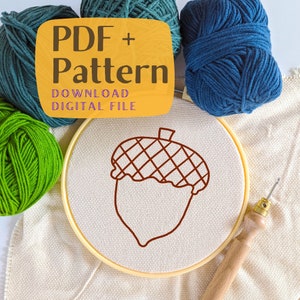 Könnte beinhalten: Eine braune Linienzeichnung einer Eichel auf einem weißen Stickrahmen. Der Rahmen ist von Garnknäuel in Grün und Blau umgeben. Der Text "PDF + Pattern DOWNLOAD DIGITAL FILE" befindet sich auf einem gelben Rechteck.