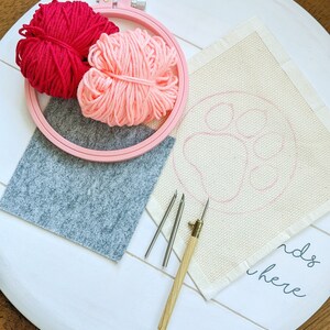 May include: A white embroidery hoop with pink and red yarn balls, three embroidery needles, a gray felt pad, and a white fabric with the words "ends here" and a pink outline of a paw print.