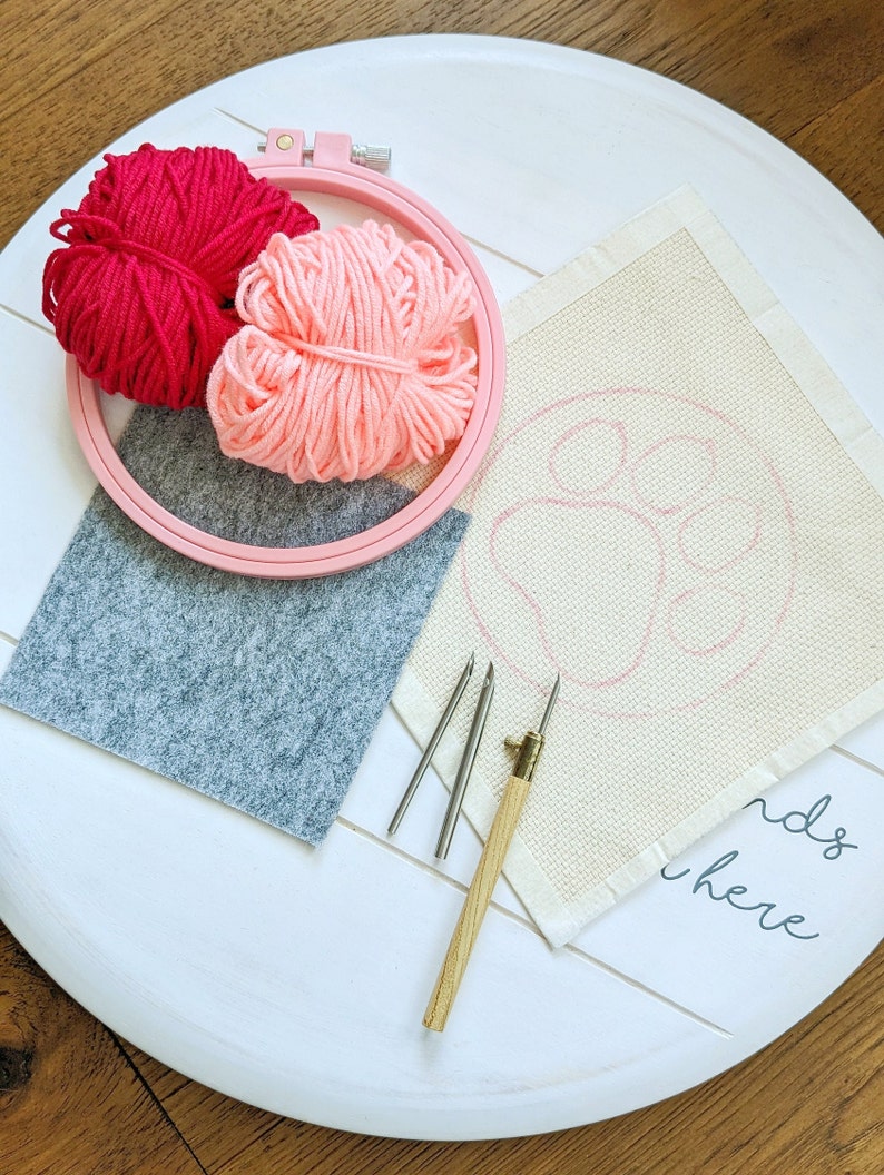 May include: A white embroidery hoop with pink and red yarn balls, three embroidery needles, a gray felt pad, and a white fabric with the words "ends here" and a pink outline of a paw print.
