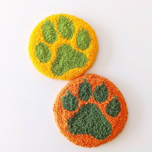 May include: Two round, plush, orange and green paw print coasters. The paw prints are green and the background is orange.