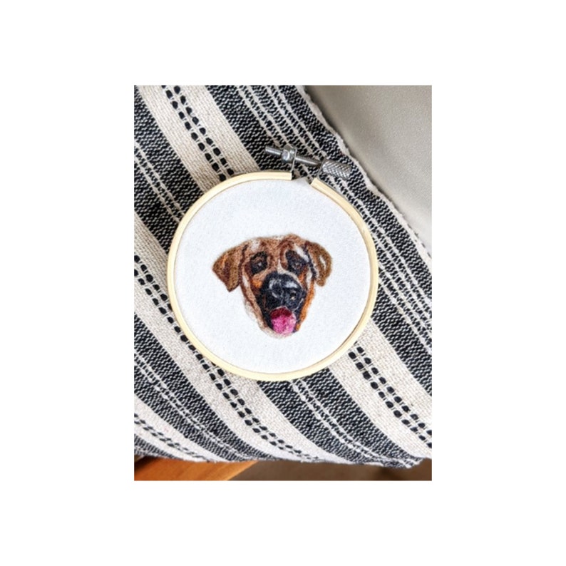 Puede incluir: La cara de un perro marr&oacute;n y beige est&aacute; bordada en una tela blanca estirada sobre un aro de bordado de madera.