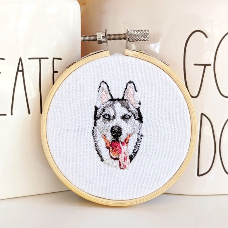 Puede incluir: Retrato bordado de un perro husky con ojos azules y la lengua rosa que sobresale, enmarcado en un aro de madera redondo.