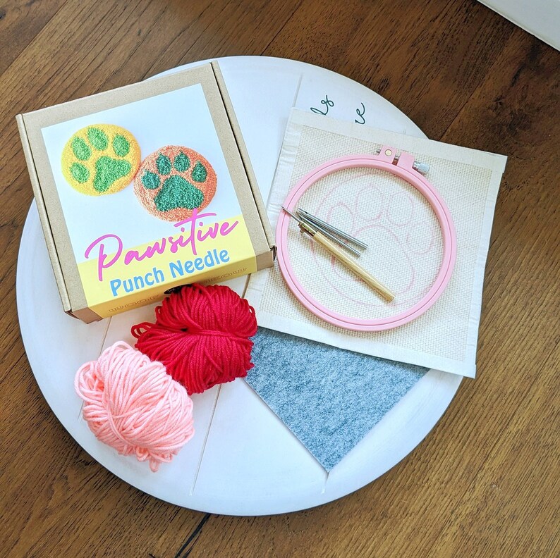 May include: A Pawsitive Punch Needle kit with two paw print designs, pink yarn, red yarn, a wooden embroidery hoop, and a needle. The kit is on a white surface.