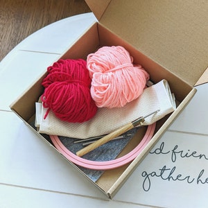 May include: A pink embroidery hoop with two balls of yarn, one pink and one red, a needle, and a piece of white fabric in a brown cardboard box. The box is open and the contents are visible. The text "and friends gather here" is written on the surface below the box.