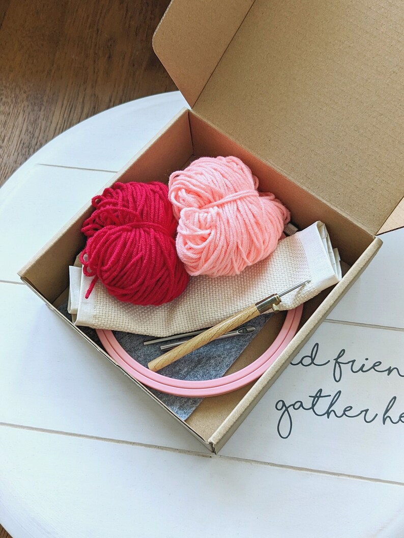 May include: A pink embroidery hoop with two balls of yarn, one pink and one red, a needle, and a piece of white fabric in a brown cardboard box. The box is open and the contents are visible. The text "and friends gather here" is written on the surface below the box.