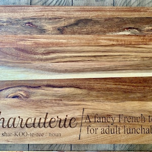 May include: A wooden cutting board with the text "Charcuterie" engraved on it. The text also includes the pronunciation and definition: "shar-KOO-te-ree / noun A fancy French term for adult lunchable."
