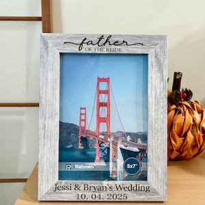 Puede incluir: Un marco de fotos de madera gris desgastada que contiene una foto de 5x7 pulgadas del Golden Gate Bridge. El marco tiene la inscripción "Father of the Bride" en la parte superior y "Jessi & Bryan's Wedding 10.04.2025" en la inferior.