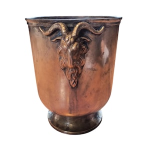 Puede incluir: Un cubo de metal color cobre con un emblema de cabeza de cabra. El cubo mide 24 cm de alto y 24 cm de ancho. La cabeza de cabra es dorada y está centrada en la parte delantera del cubo. El cubo tiene una base de pedestal.