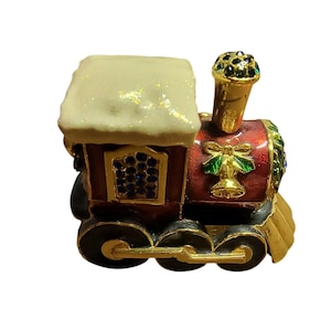 May include: A red and gold toy train with a white snow-covered roof. The train has a bell and holly accents.