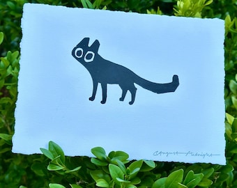 May include: A rectangular piece of white paper with deckled edges featuring a black ink illustration of a stylized cat with large circular eyes. The text August Midnight is written in black ink in the bottom right corner of the paper.