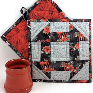 May include: A red ceramic mug and two quilted Christmas-themed pot holders. One pot holder is square with a black background and red poinsettias, the other is triangular with a red background and a black pattern. Both have hanging loops.