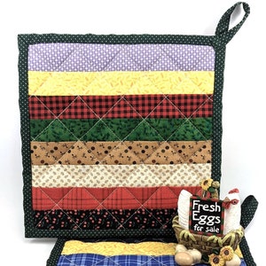 Puede incluir: Dos agarraderas de olla acolchadas con un tema de granja. La agarradera superior tiene un borde verde con un patrón de rayas morado, amarillo, rojo, verde, marrón, rojo y negro. La agarradera inferior tiene un borde verde con un patrón de rayas azul, rojo, verde, amarillo y azul. Una figurita de pollo de cerámica con un letrero que dice "Fresh Eggs for Sale" está sentada en la agarradera inferior.