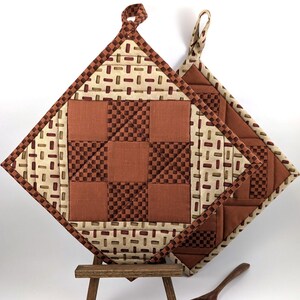 May include: Two quilted pot holders with a brown, tan, and cream color scheme. The pot holders have a square design with a checkered pattern and a geometric pattern. The pot holders are displayed on a wooden easel.