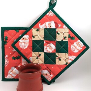 Pode incluir: Dois pegadores de panela acolchoados vermelho e verde com um padrão de Papai Noel. Os pegadores têm um laço para pendurar. Uma caneca de cerâmica vermelha está em primeiro plano.
